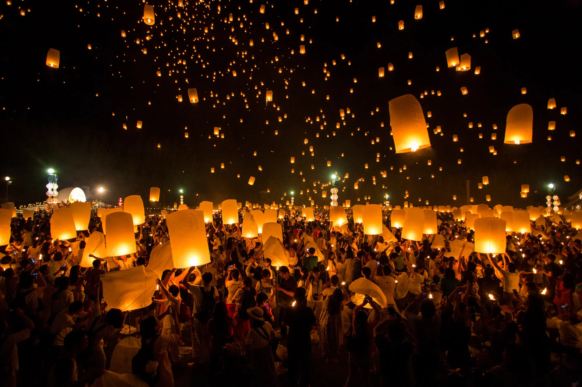 Thousands of glowing paper lanterns released into the night sky during Loi Krathong festival, with crowds below holding lit lanterns.
