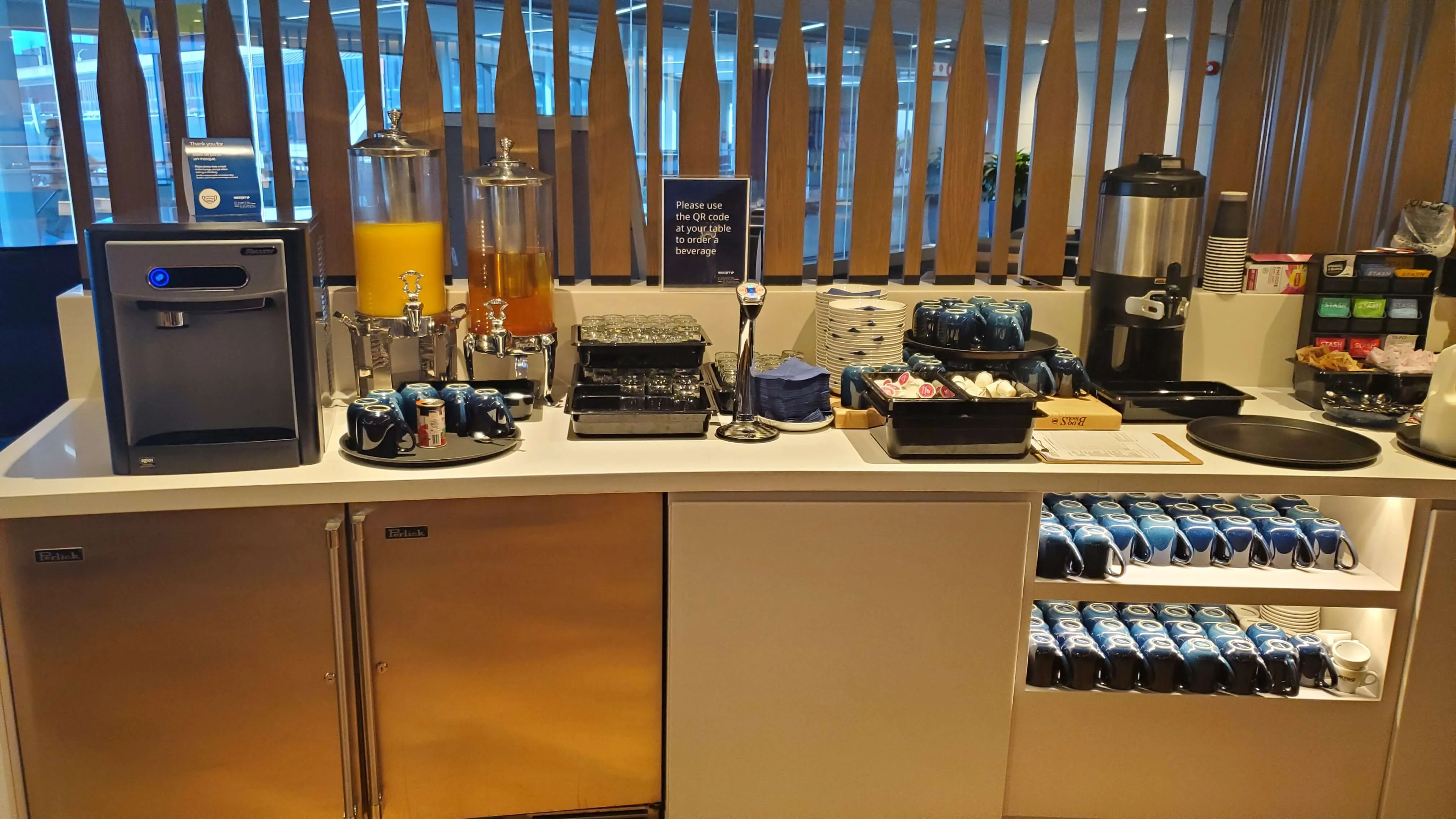 Airport lounge beverage and snack station with coffee maker, juice dispenser, pastries, and packaged items on white counter with wood slat backdrop.