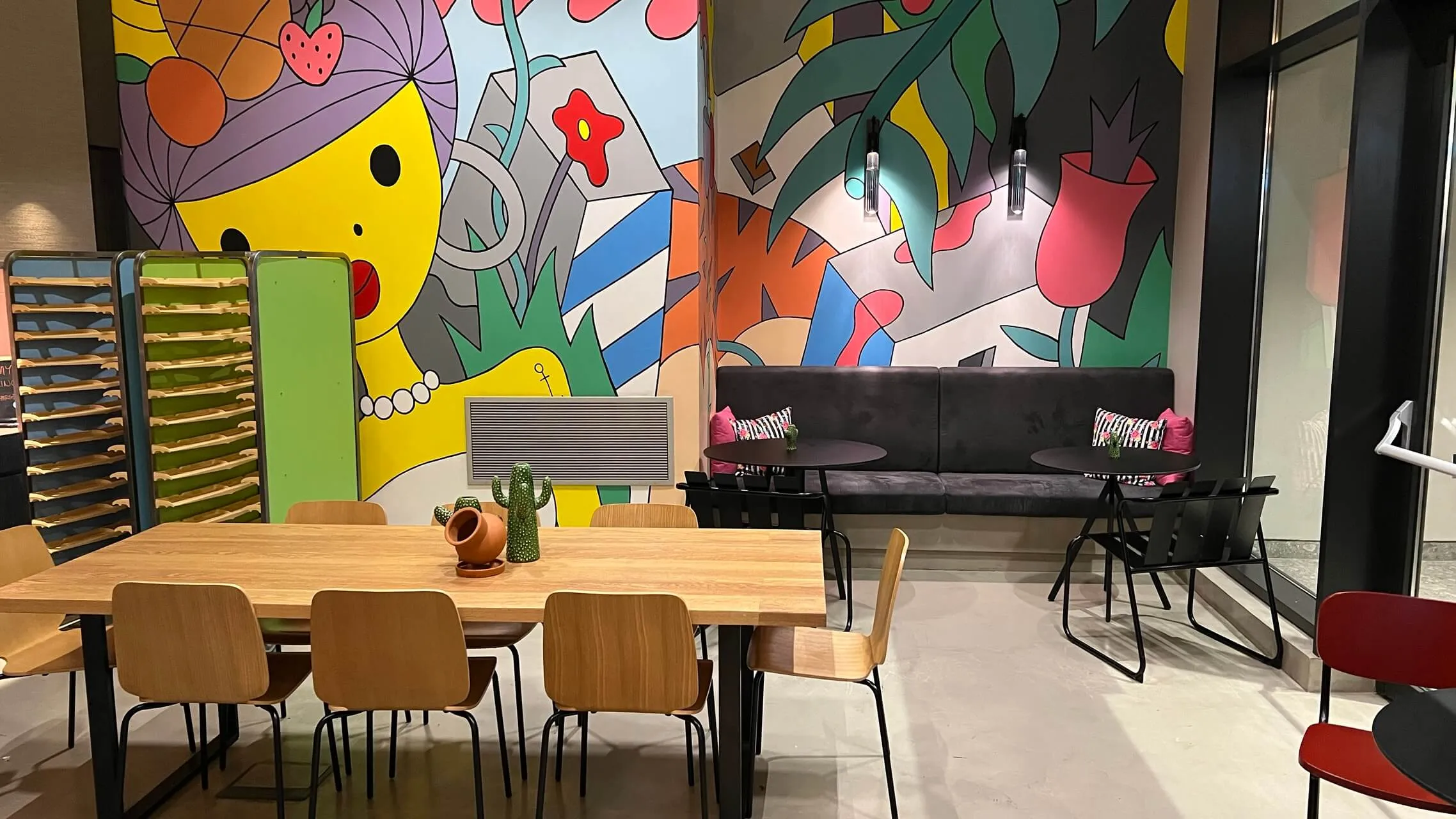 Colorful lounge seating area with wooden table, tan chairs, and vibrant pop-art wall murals featuring yellow faces and flowers