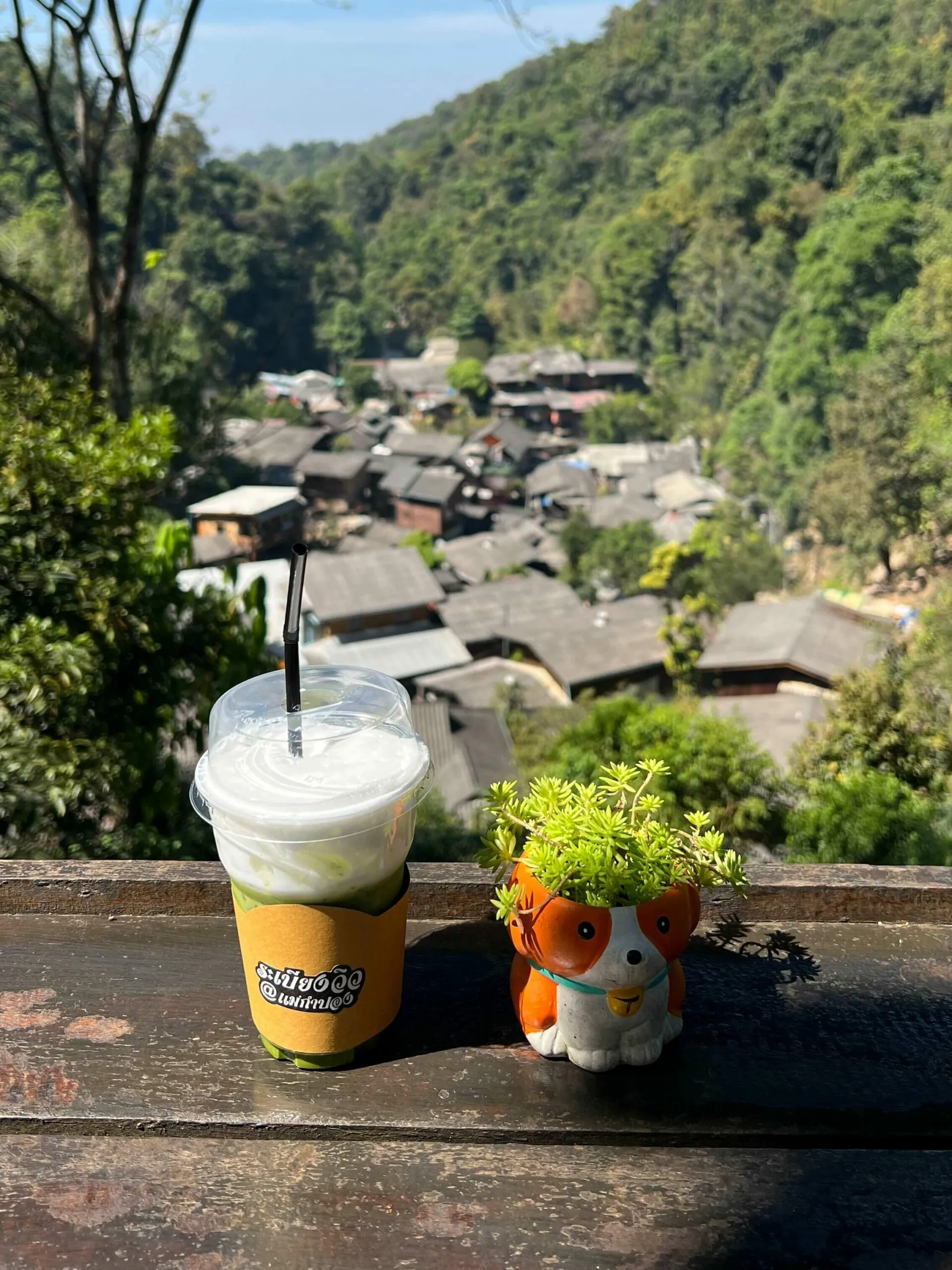 Iced matcha latte with yellow sleeve and lucky cat figurine on wooden railing overlooking forested Mae Kampong village with traditional dark-roofed buildings nestled in green hillside.