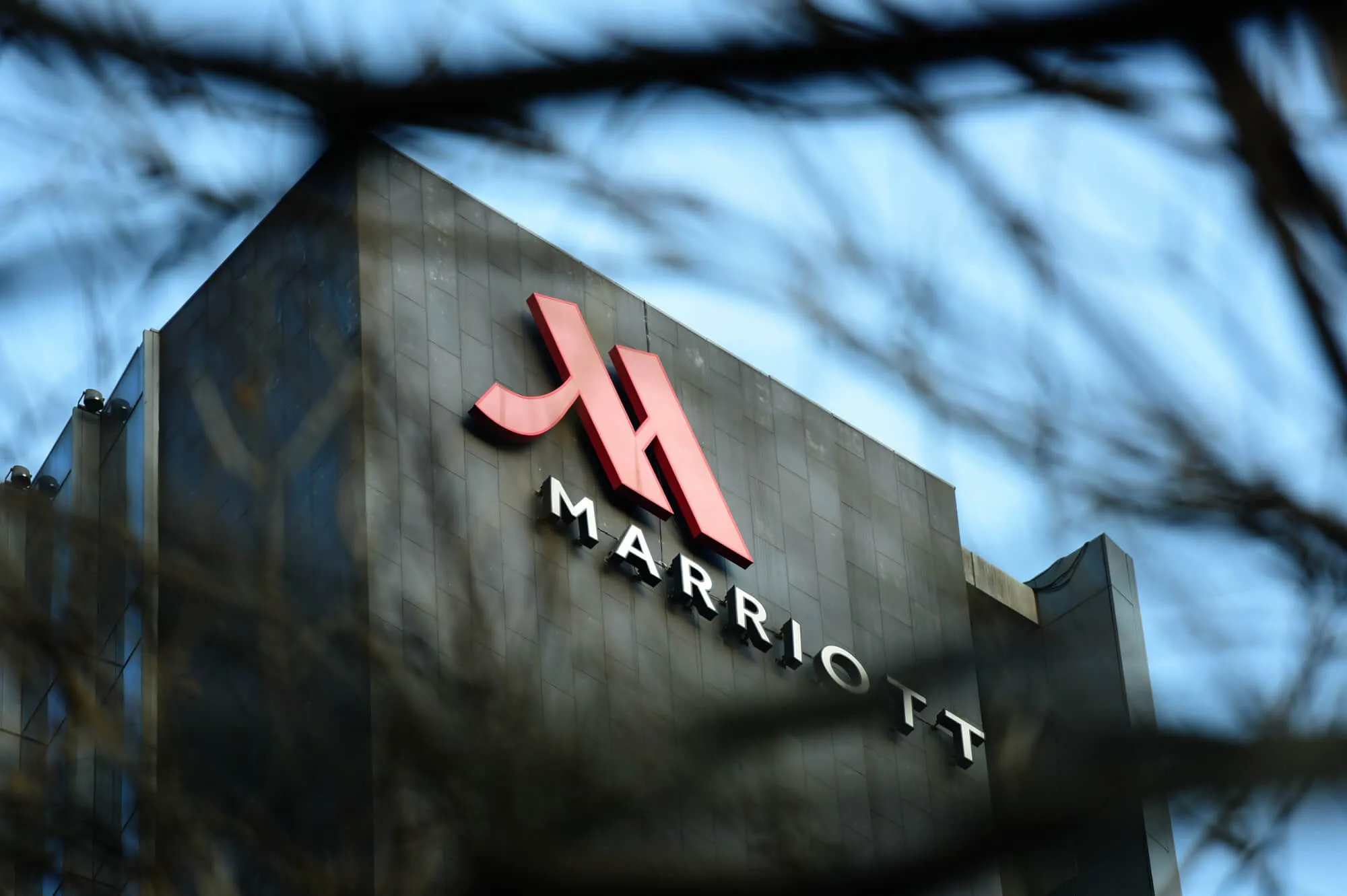 Marriott red chevron logo and text on dark exterior building signage with tree branches overhead
