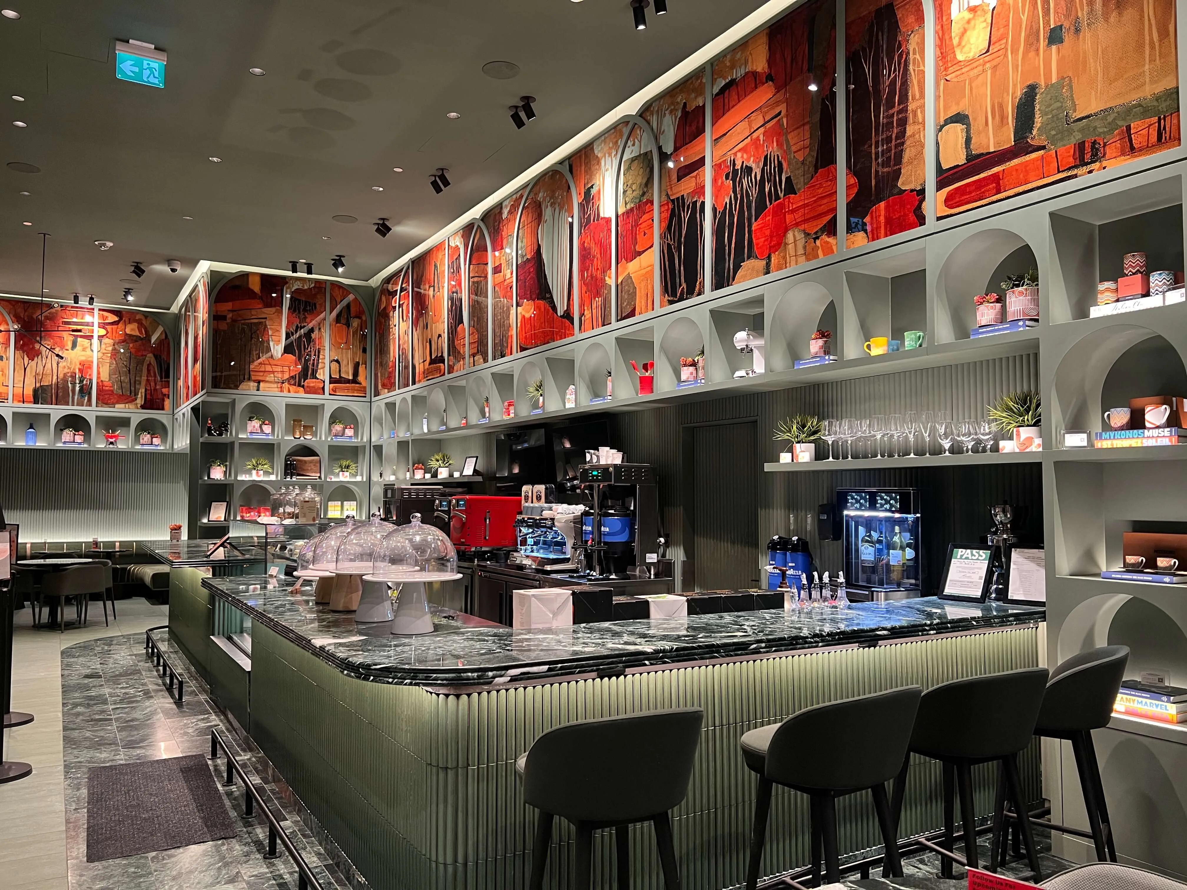 Modern cafe interior with green-paneled counter, black bar stools, marble countertop, white shelving, and red artistic panels on upper walls.