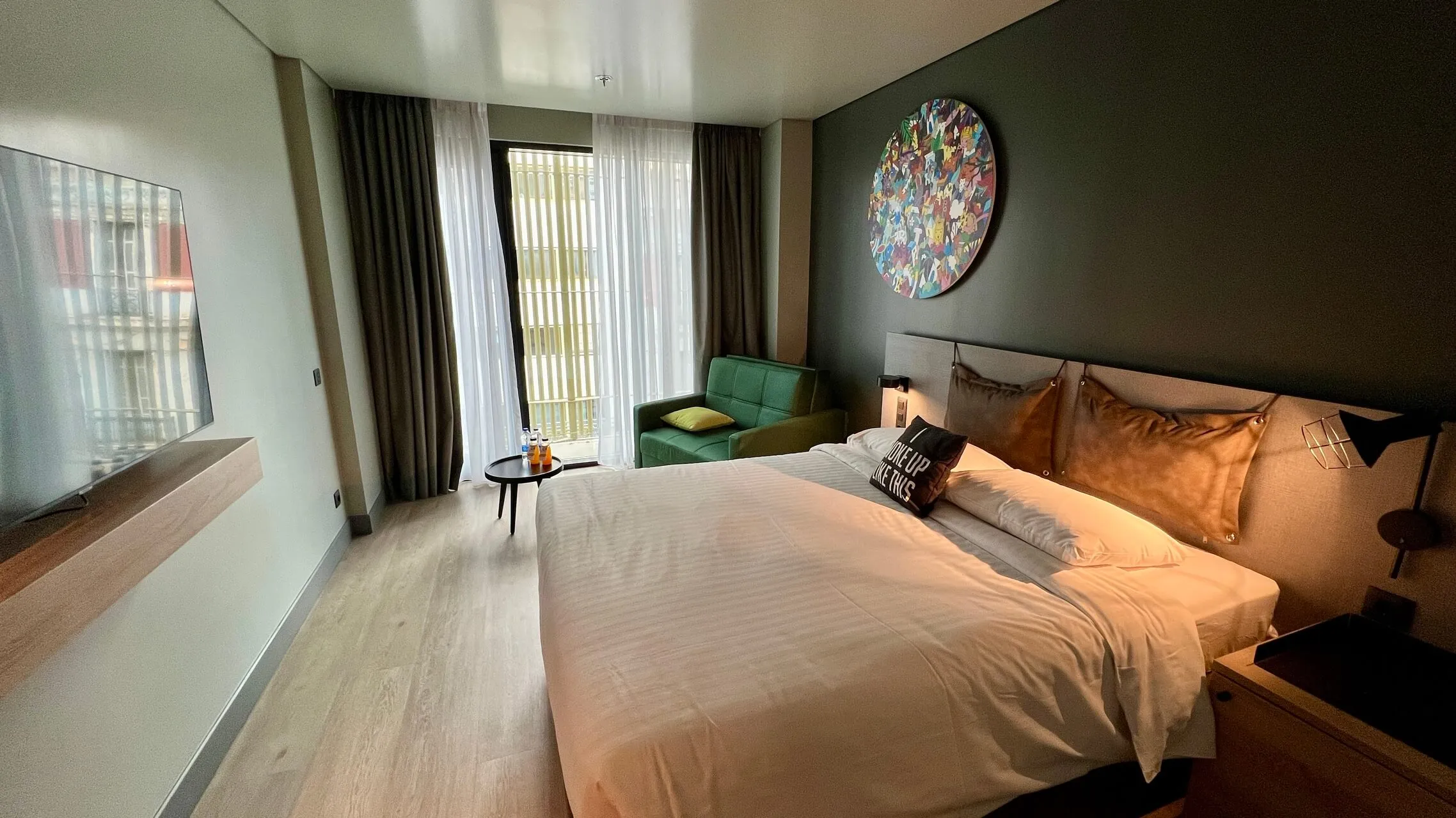 Modern hotel bedroom with king bed, copper pillows, dark accent wall, green armchair, floor-to-ceiling windows with sheer and blackout curtains, and colorful wall art.