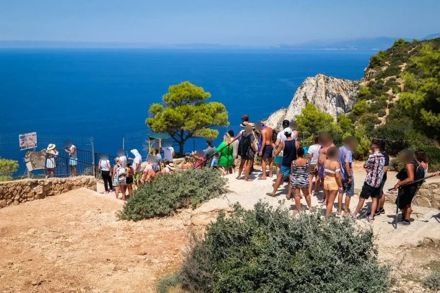 Tourists at a clifftop viewpoint overlooking blue Mediterranean Sea with rocky coastline and pine trees, Navagio Beach area Greece