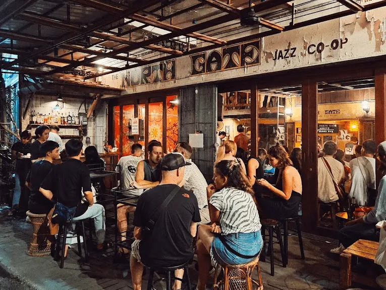 Crowded jazz bar exterior at night with patrons seated outside and standing inside storefront, neon signage visible