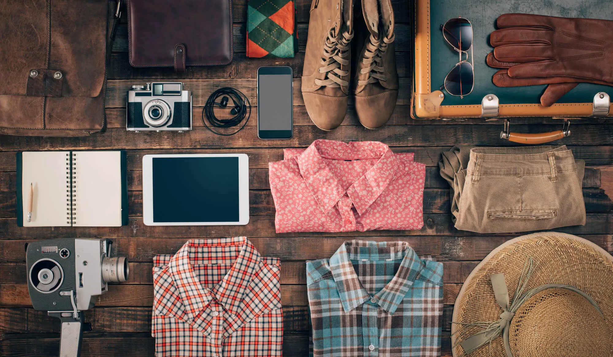 Flat lay of travel packing essentials on wood surface: leather bags, boots, camera, tablet, notebooks, plaid and checkered shirts, hats, gloves, and accessories.