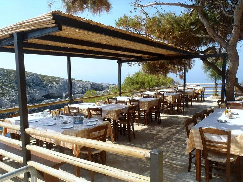 Outdoor tavern with wooden tables and chairs under pergola, Mediterranean sea and rocky coastline visible beyond, traditional Greek setting.