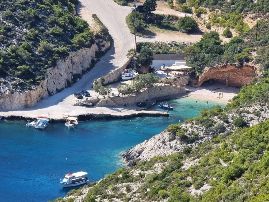 Aerial view of Porto Vromi beach cove with turquoise water, white sand, moored boats, and limestone cliffs surrounded by pine trees
