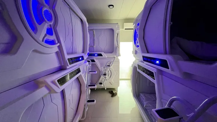 Purple-lit capsule pod beds with individual screens and storage in a modern hostel corridor.
