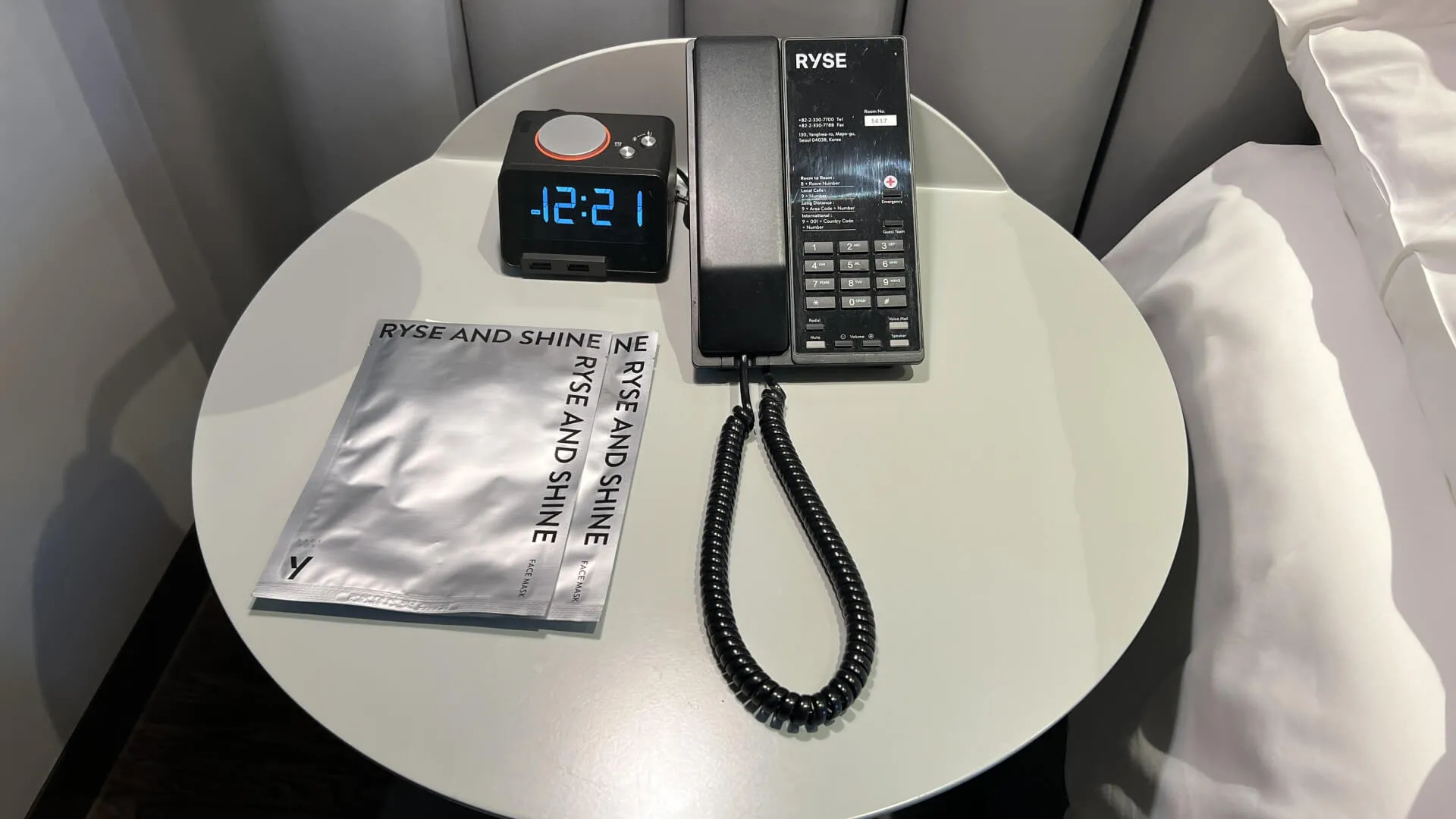RYSE hotel nightstand with black desk phone, blue digital alarm clock showing 4:21, and amenity packets