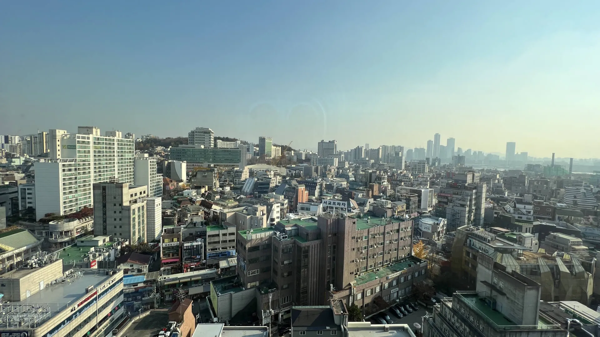 Aerial cityscape from high-rise hotel room showing dense urban neighborhood with downtown skyline in distance under clear blue sky