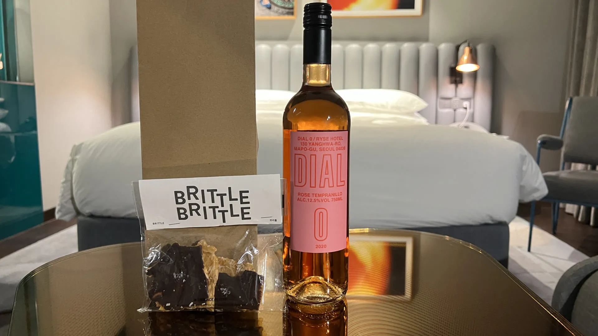 Welcome amenities on desk: Brittle Brittle candy and Dial rosé wine bottle in modern hotel room with bed in background