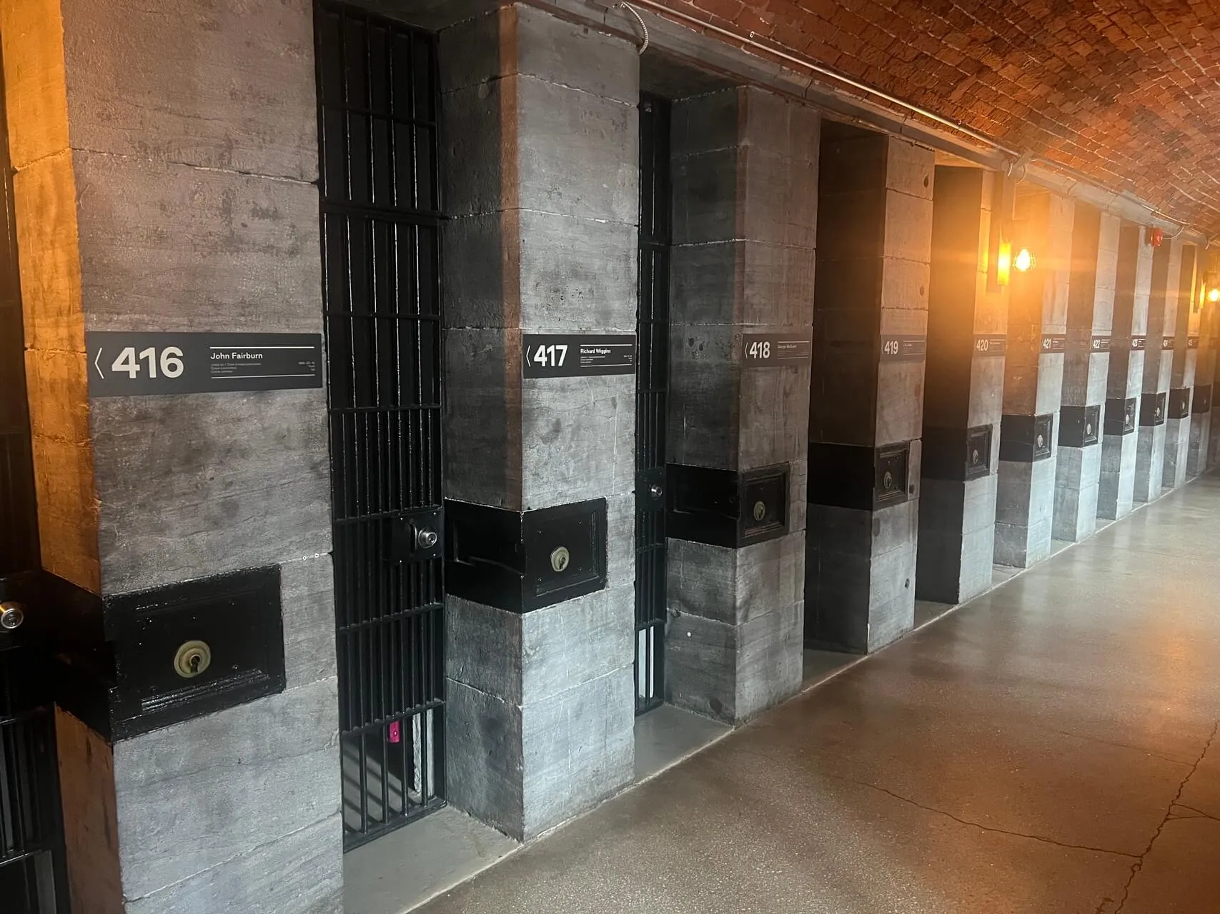 Hotel corridor with numbered room doors (416, 417, 418) along concrete wall with exposed brick ceiling and warm lighting.