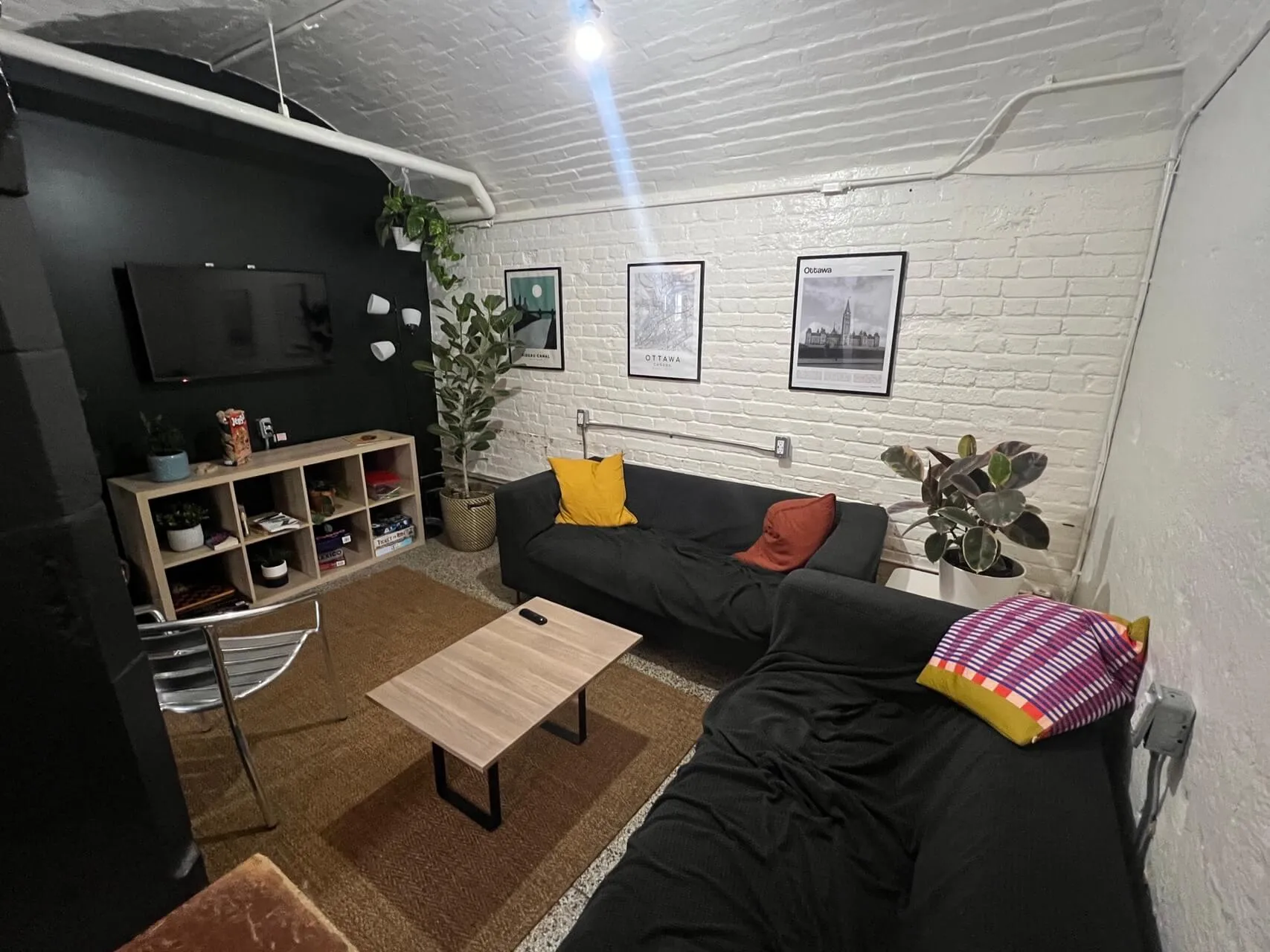 Basement lounge with black sofa, wood coffee table, shelving unit, wall-mounted TV, framed city prints, and potted plants