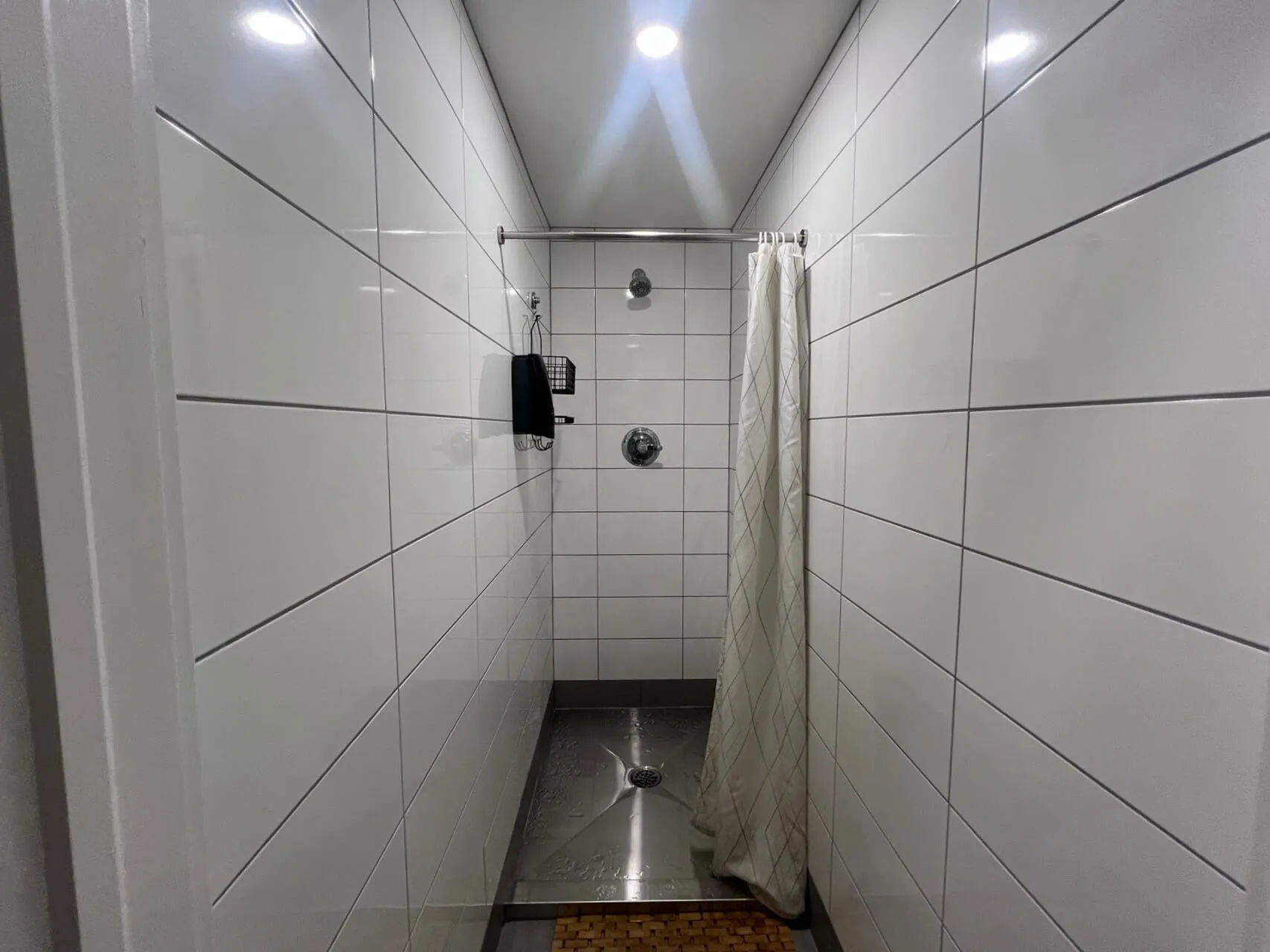 Narrow shower stall with white subway tiles, dark grout lines, chrome fixtures, and white shower curtain in a compact bathroom