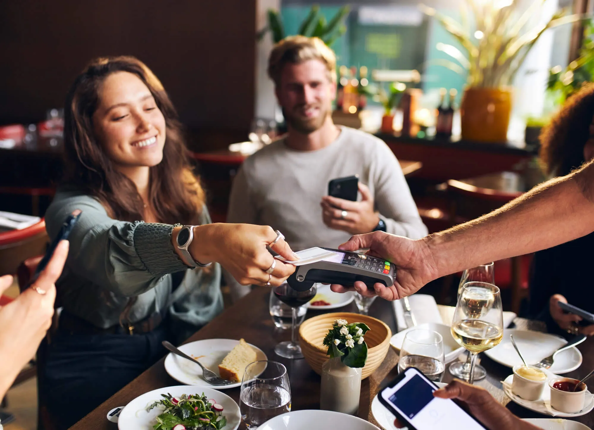 Woman paying with contactless credit card at restaurant dinner table with friends and white wine.