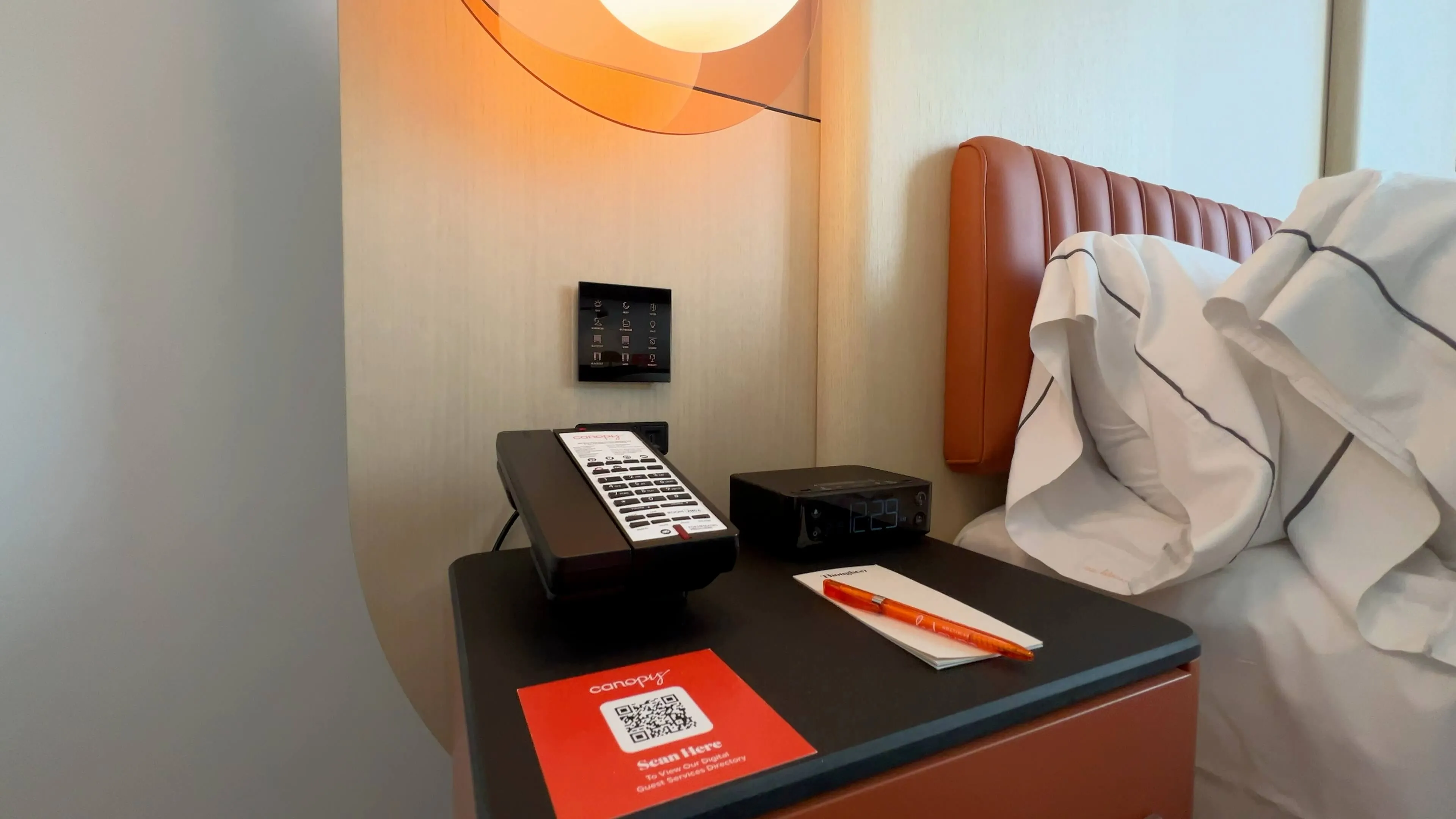 Bedside table at Canopy by Hilton Toronto Yorkville with remote, orange accent headboard, and warm lighting