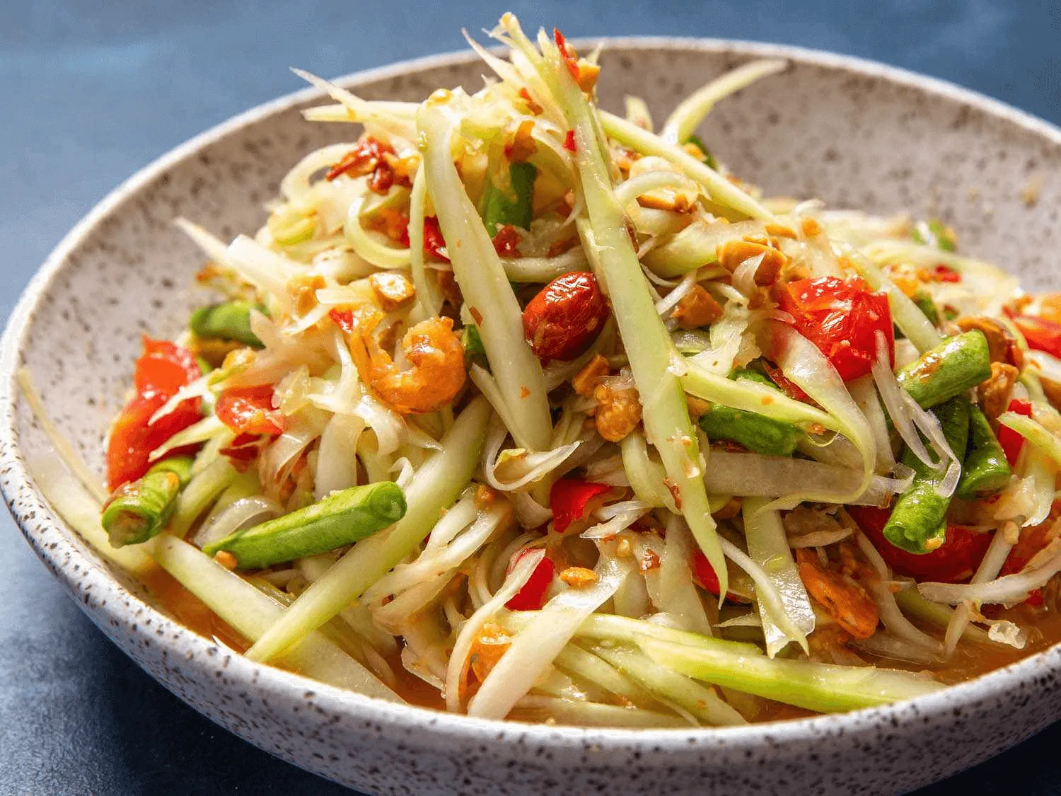 Som tam papaya salad with shredded papaya, green beans, peanuts, and red chilies in a gray ceramic bowl