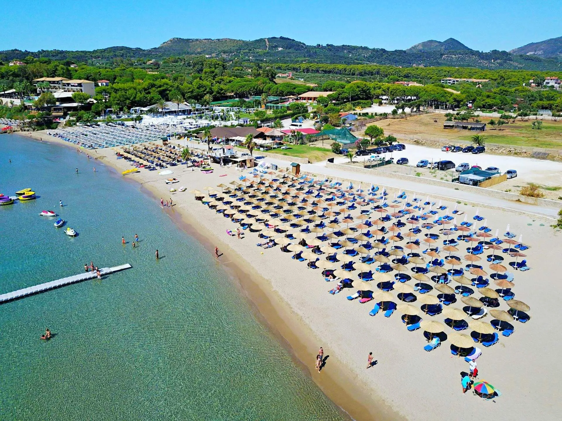 Aerial view of St. Nicholas Beach with organized blue and black umbrellas, clear turquoise water, beach facilities, and green mountains in the background.