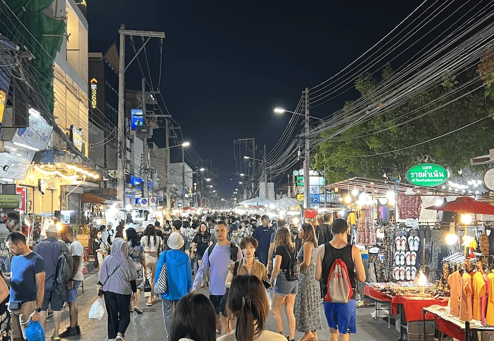 Crowded night market street in Bangkok with vendor stalls, tangled power lines, and neon signs.