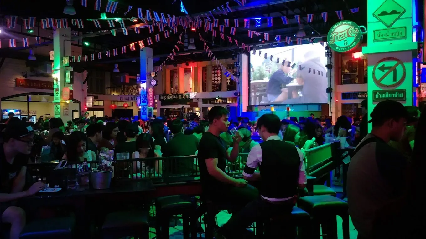 Crowded Bangkok street food market at night with neon green and purple lighting, vendor stalls, and large screen displaying content.