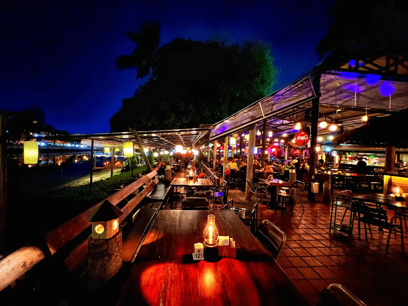 The Goodview Bar's outdoor waterfront terrace at night with colored lantern lighting, wooden tables, and diners under a pergola structure
