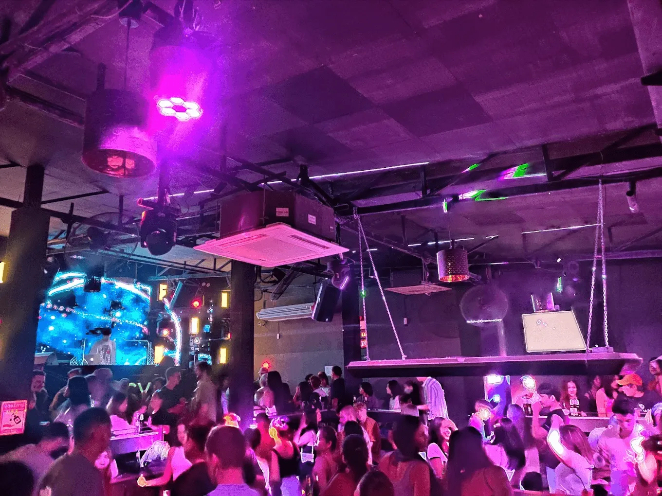 Nightclub interior with purple and pink neon lighting, elevated DJ booth, and crowd of people dancing