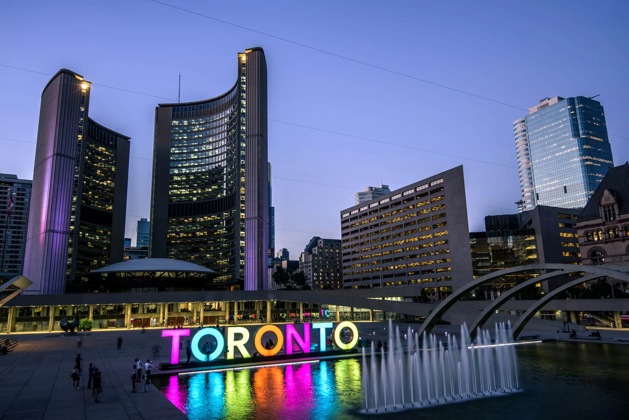 Colorful TORONTO sign at Nathan Phillips Square at dusk, with Toronto skyline and city hall reflected in fountain pool