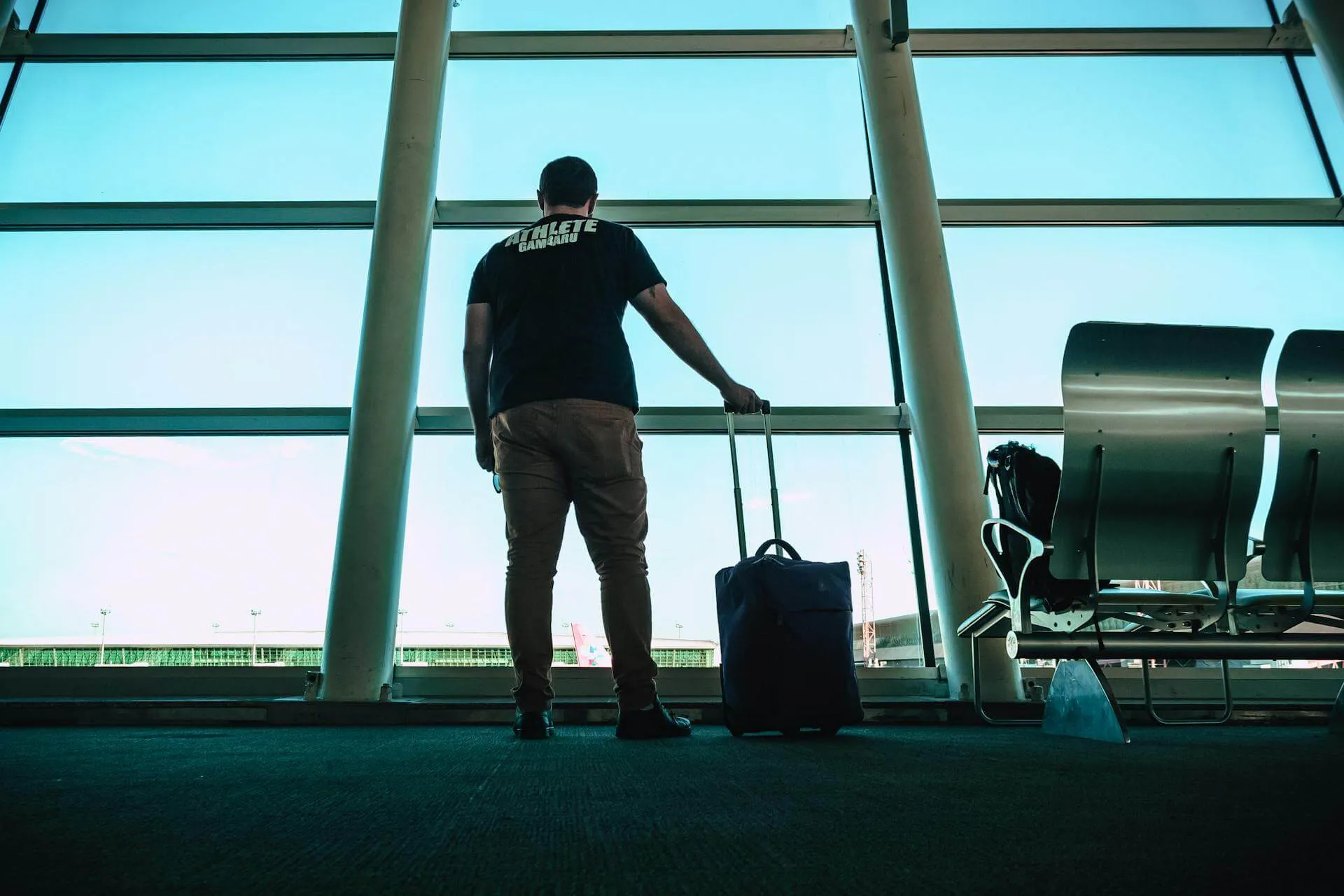 Traveler with rolling suitcase standing at airport window with bright sky view and seating area behind.