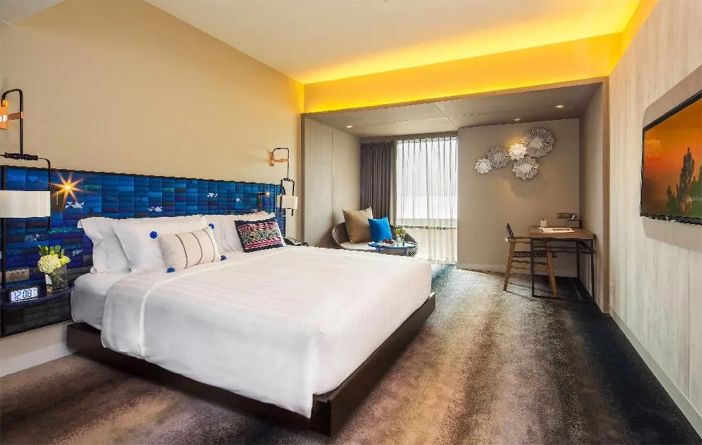 Modern hotel room with king bed, blue tile headboard, yellow ceiling accent lighting, and separate seating alcove with desk at U Nimman Chiang Mai