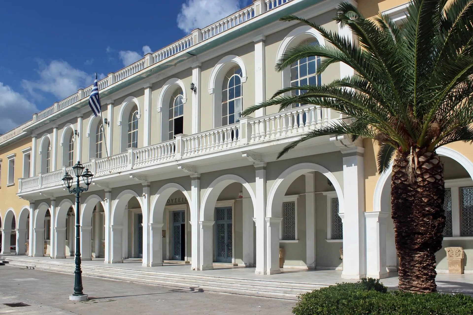 White neoclassical building with arched colonnade, palm tree, and blue sky in Zakynthos Town
