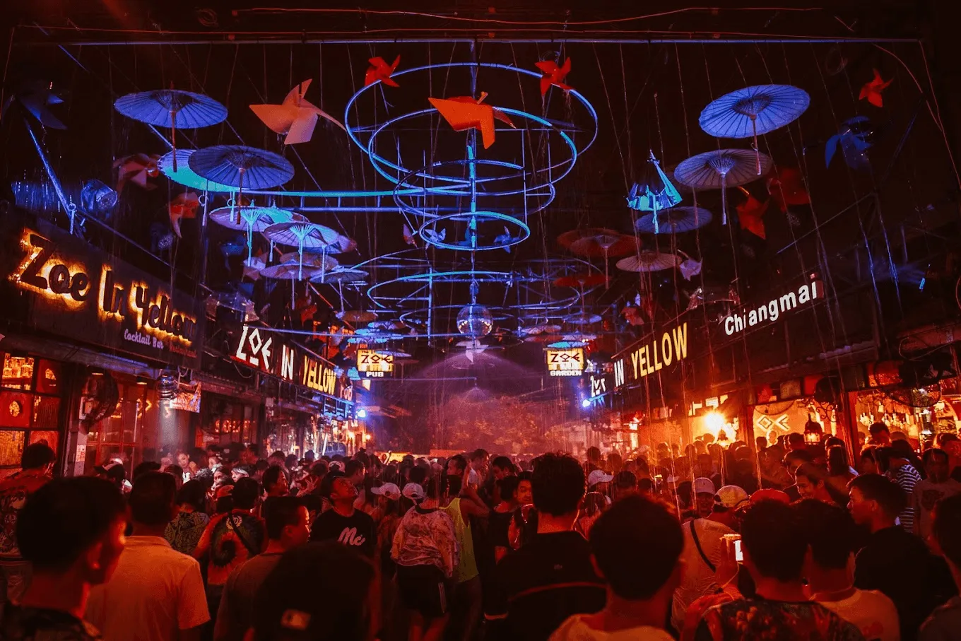 Crowded night market street in Chiang Mai with neon signs, red lantern decorations, and illuminated umbrella installations overhead.